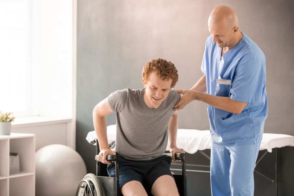 Clinician in uniform helping young man in pain to sit in wheelchair