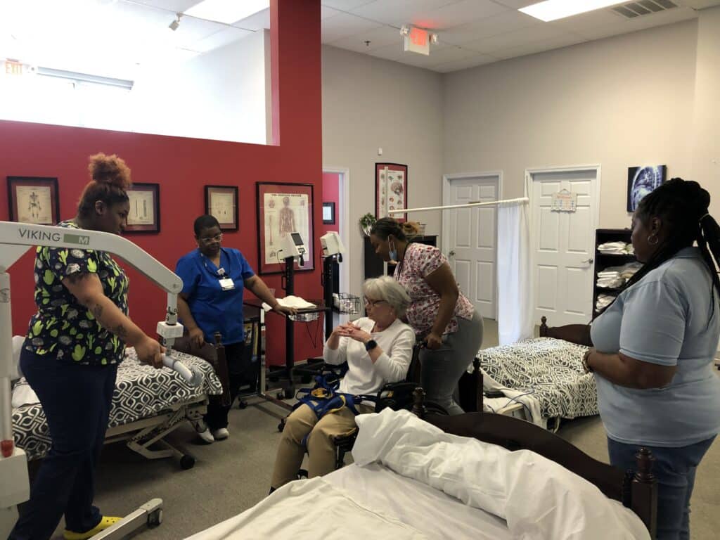 Students and Ambassadors listen to instructor Nora Gillespie, seated in a wheelchair in the center, as they learn to lift her back into the bed using a large piece of equipment.