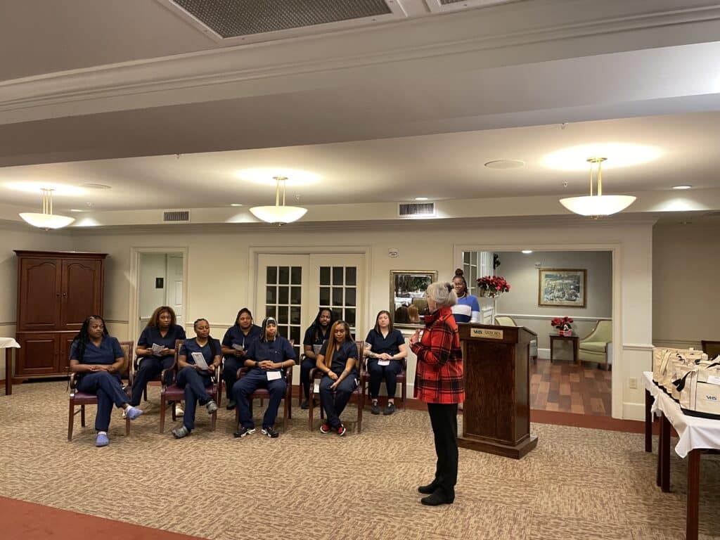 Instructor Nora Gillespie addresses the class of eight graduates, seated, wearing navy blue scrubs.